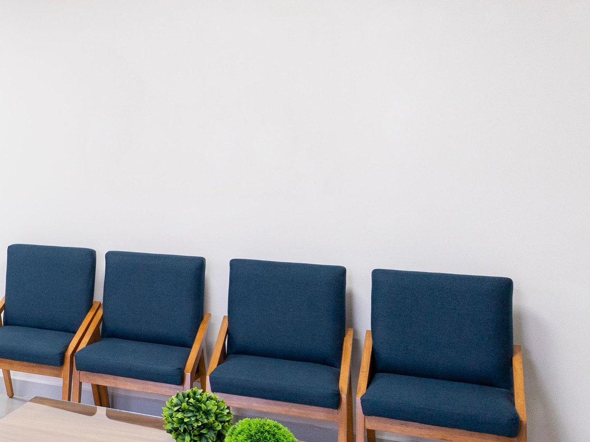 Quatro poltronas azuis vazias, lado a lado, numa sala de espera clara